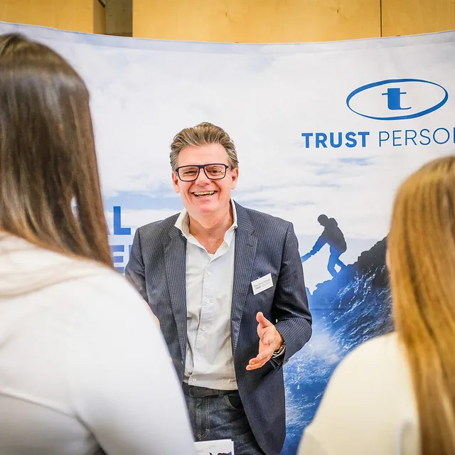 Herr Mag. Roman Ettinger, Geschäftsführer von TRUST Personal spricht lächelnd mit zwei Besucherinnen an einem Informationsstand in der HAK Steyr.