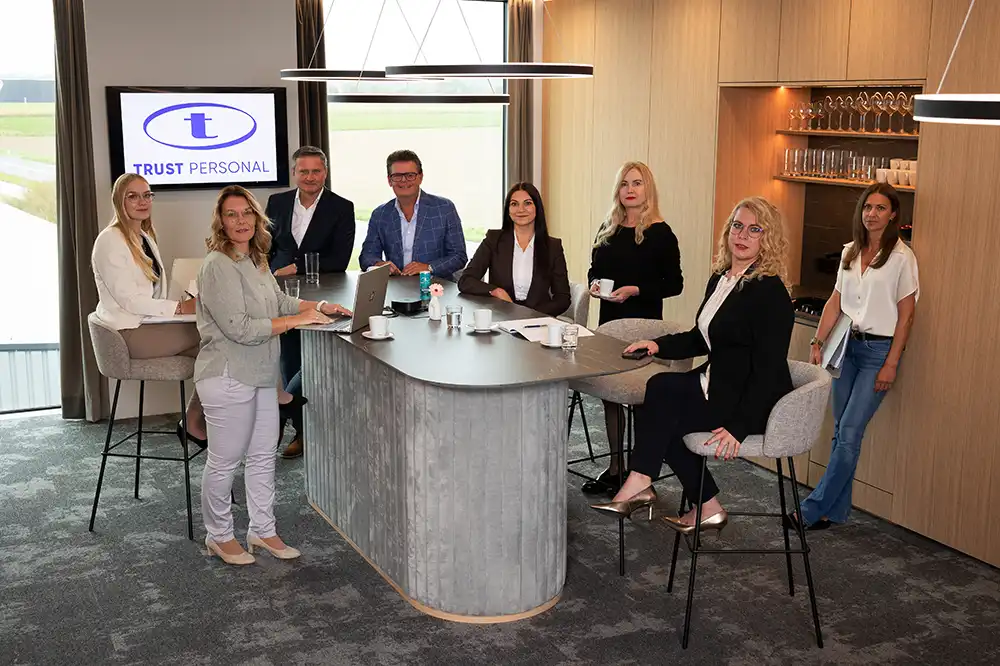 Gruppenbild des TRUST Personal Teams um den großen Stehtisch in der modernen Office-Lounge.