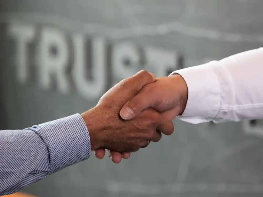 Nahaufnahme von zwei Männern beim Handschlag vor einer Wand mit dem TRUST Personal Logo.